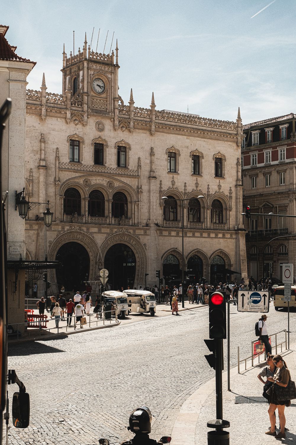 Historic Rossio train station facade with ornate architecture, clock tower, cobblestone square, and pedestrians in Lisbon, Portugal.