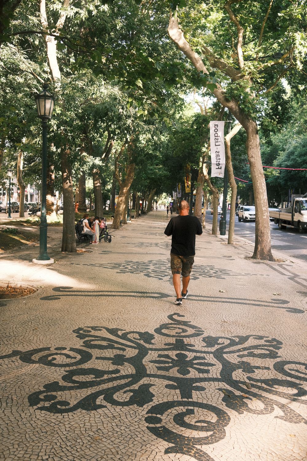 Man walking on ornate mosaic cobblestone path lined with trees and vintage lamp posts in a European city.