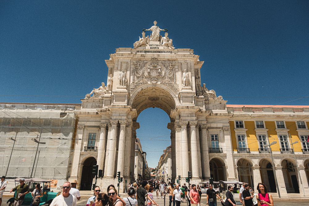Tourists gather at the iconic Rua Augusta Arch in Lisbon's Commerce Square on a sunny day.