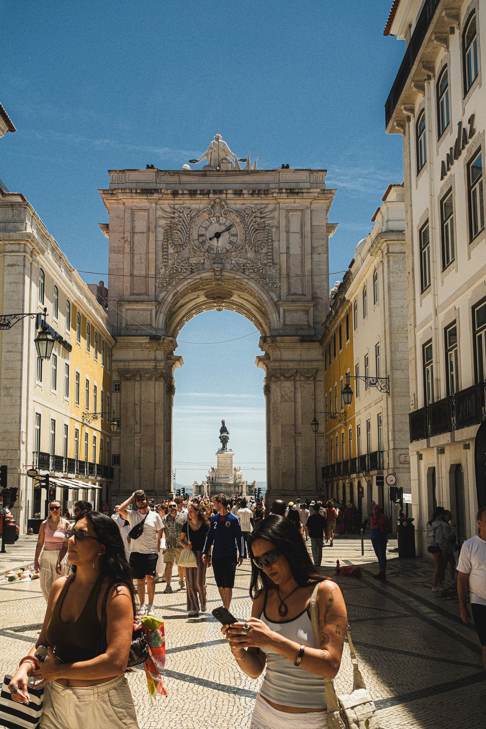 Tourists walking through Rua Augusta Arch in Lisbon, Portugal on a sunny day with blue skies.