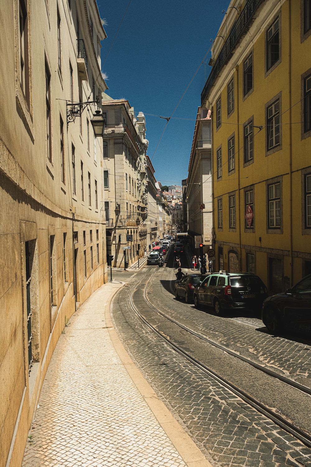Narrow cobblestone street with tram tracks in Lisbon, Portugal, flanked by yellow and beige historic buildings under blue sky.