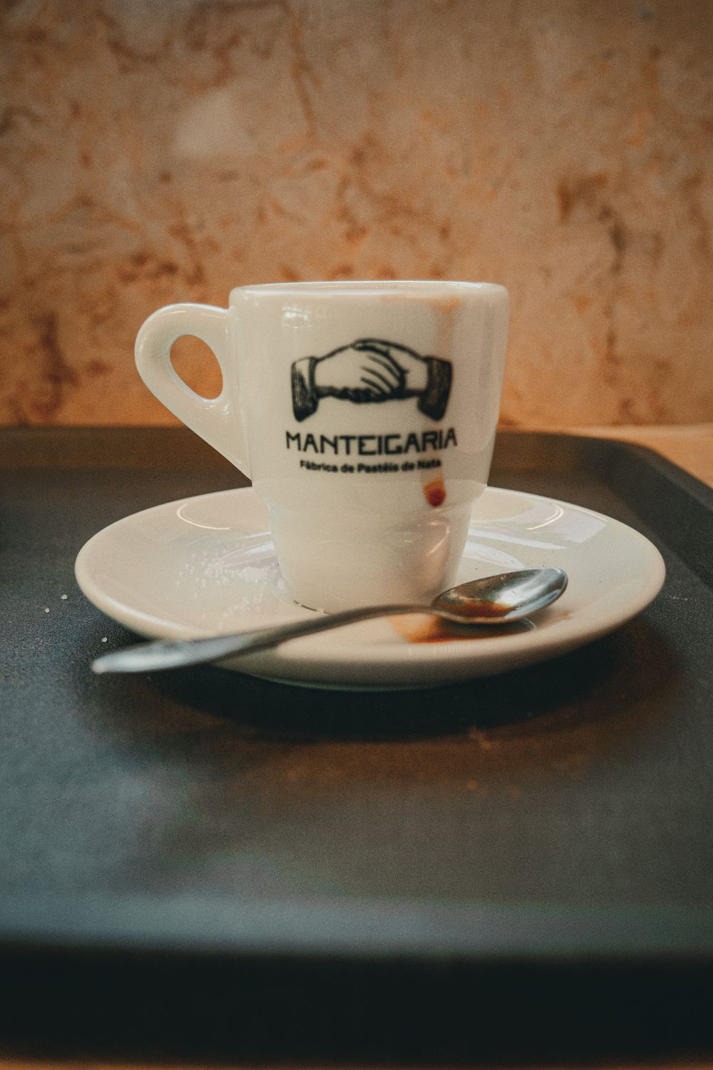 White Manteigaria espresso cup and saucer with spoon on dark tray, Fabrica de Pasteis de Nata branding.