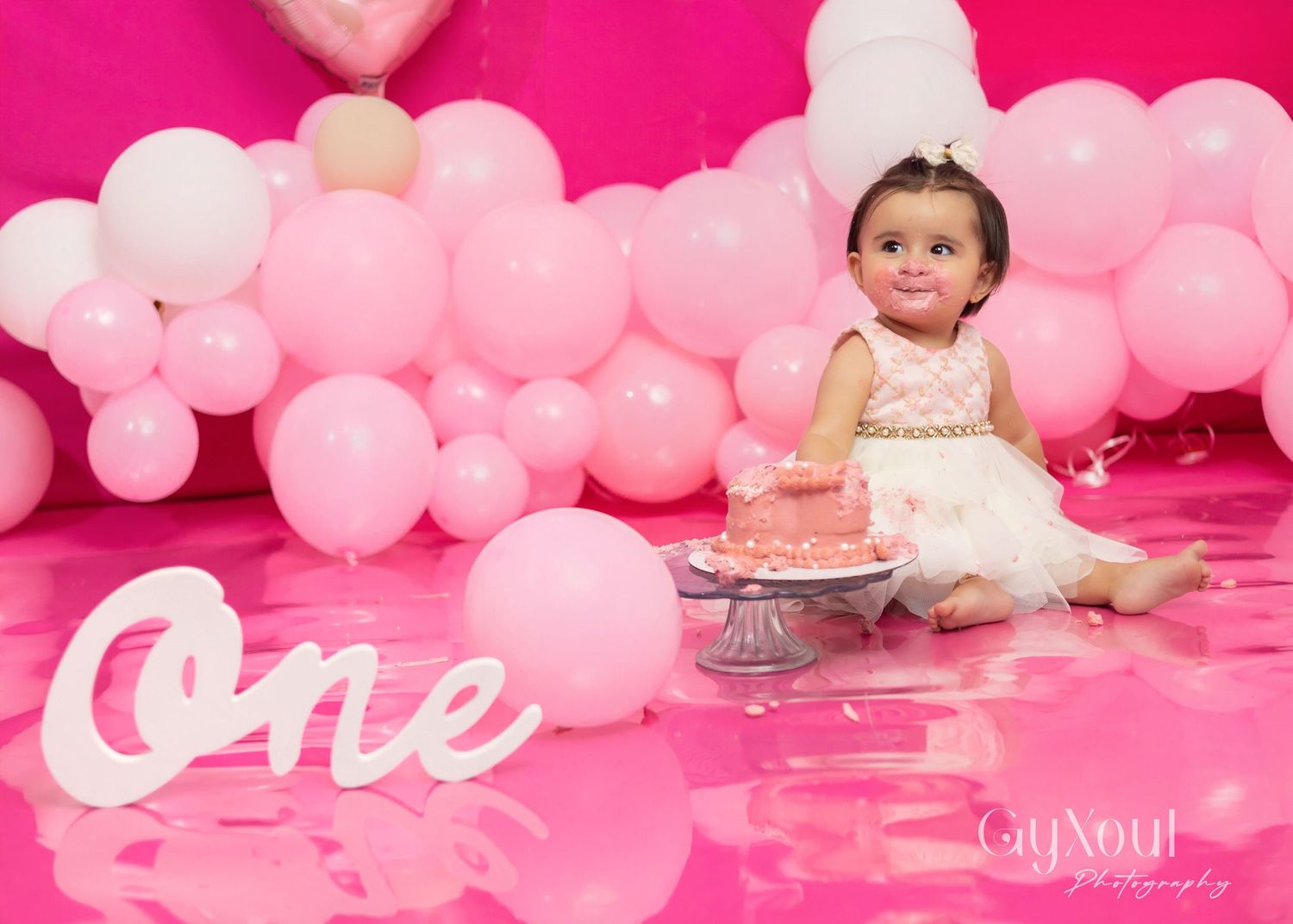 Baby girl in white dress celebrates first birthday with pink cake surrounded by pink balloons and One sign.