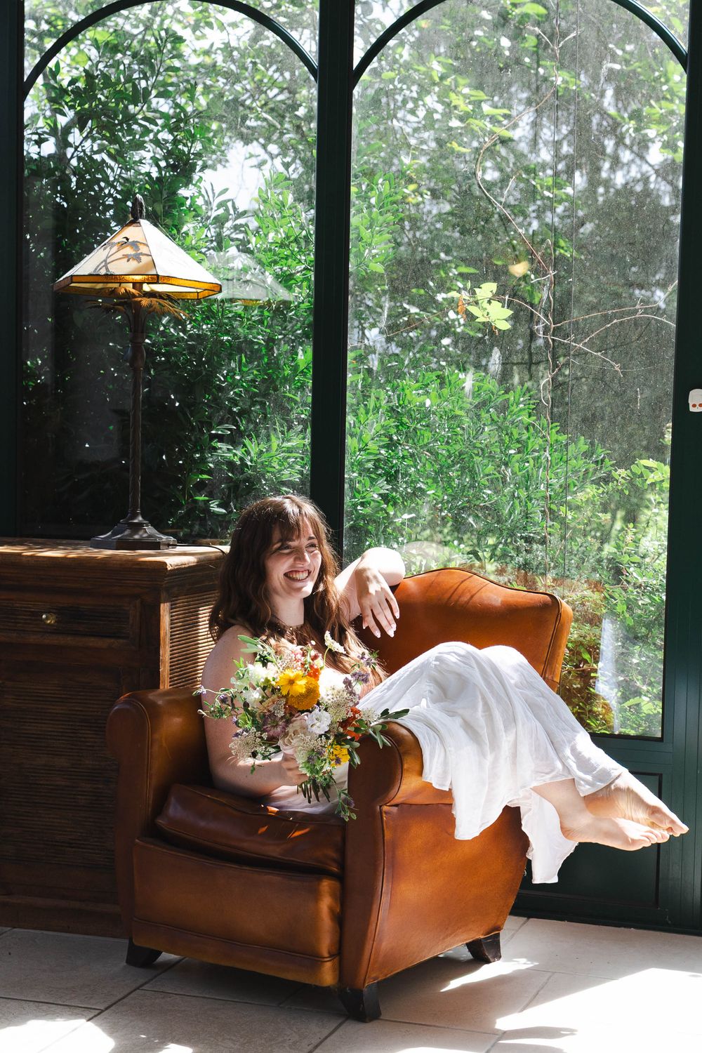 Smiling bride in white dress holding colorful bouquet, seated in brown leather armchair by sunlit greenhouse windows.