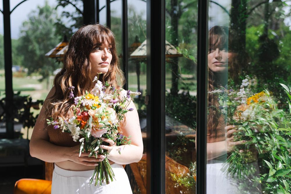 Bride in white dress holding wildflower bouquet stands by glass door with garden reflection.