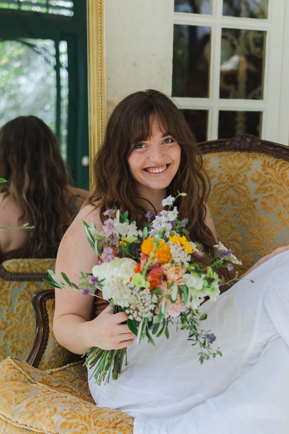 Smiling bride holding colorful wildflower bouquet seated in ornate gold chair near mirror.