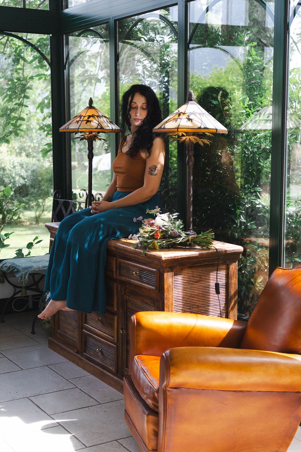 Woman in teal skirt sits on wooden cabinet flanked by Tiffany lamps in sunlit glass conservatory with leather armchair.