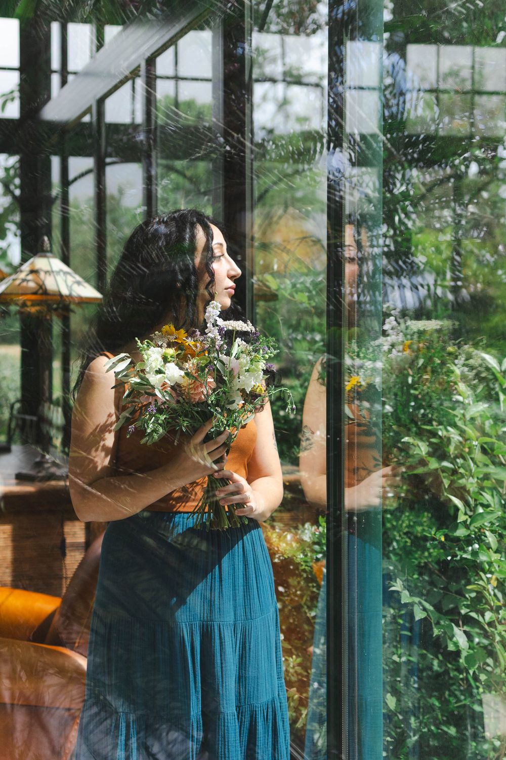 Woman in teal dress holding wildflower bouquet standing by glass greenhouse windows with lush green plants.