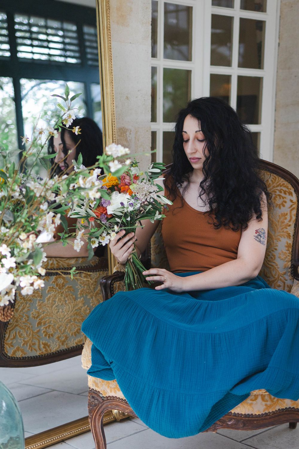 Woman in teal skirt and brown top sits in ornate chair holding a wildflower bouquet near a gilded mirror.