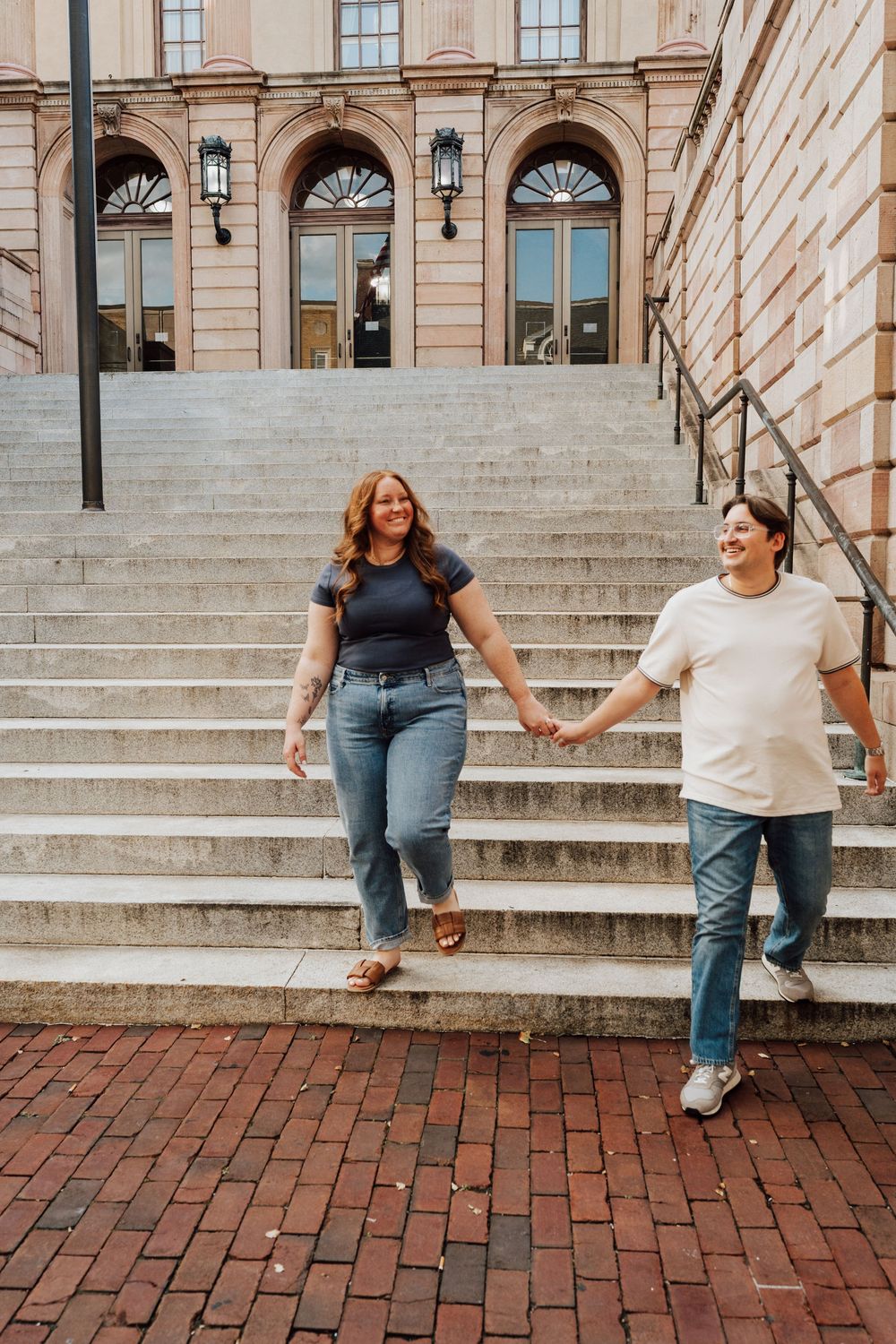 Happy couple holding hands walking down grand stone steps outside historic building on brick plaza.