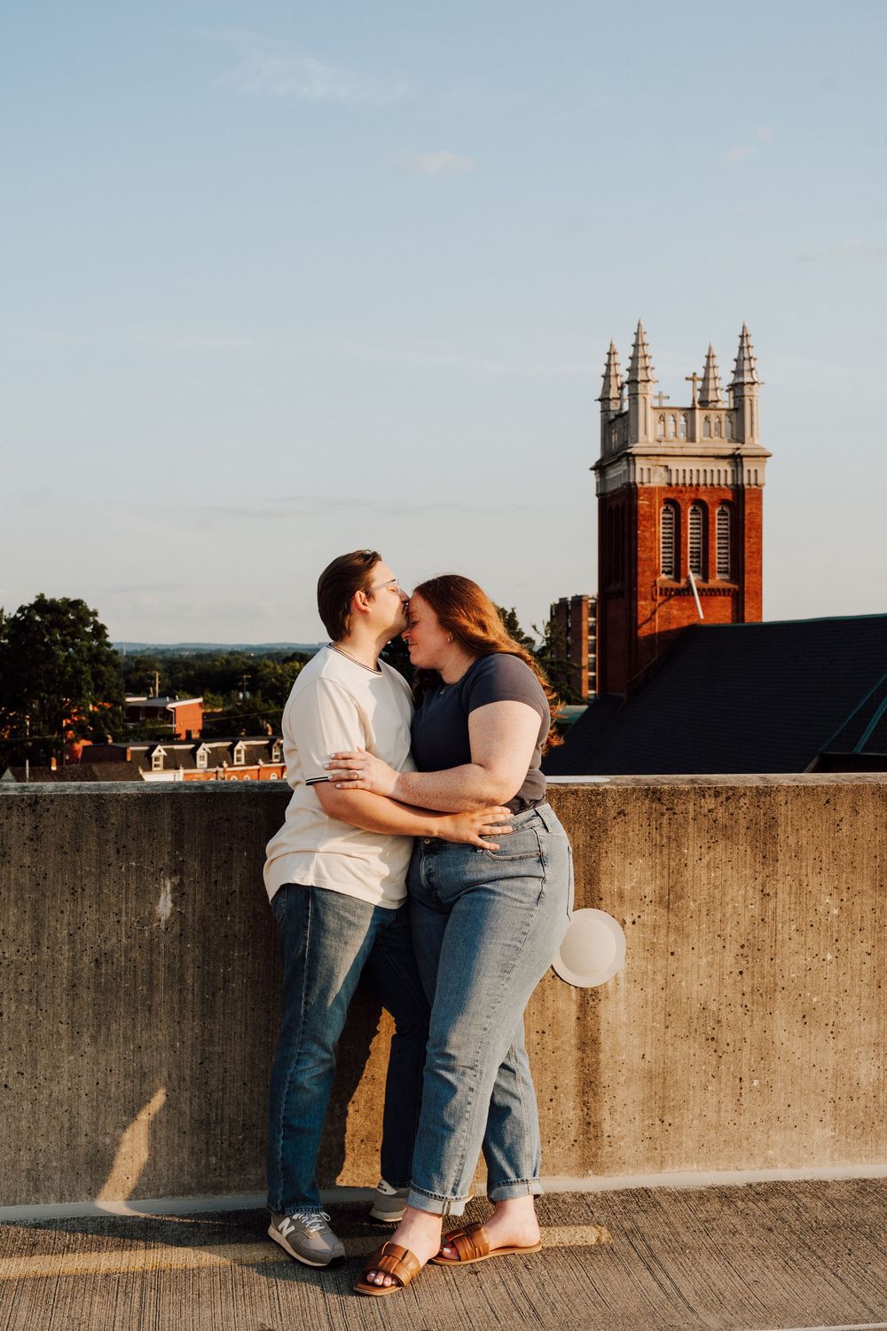 Couple sharing a romantic kiss on a rooftop at golden hour with a historic brick church tower in the background.