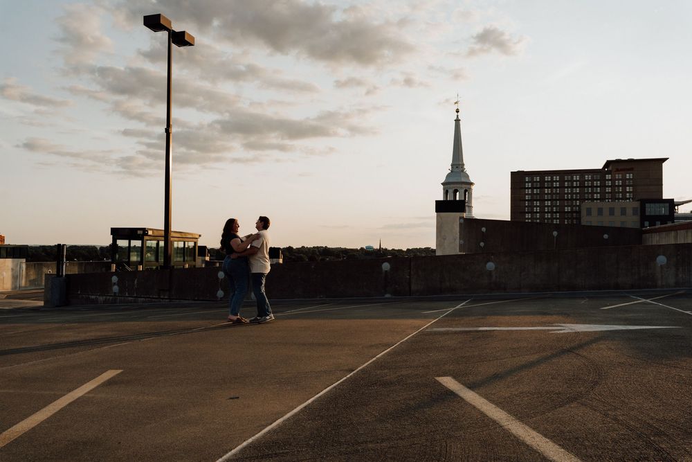 Couple dancing on rooftop parking lot at sunset with church steeple and city buildings in background.