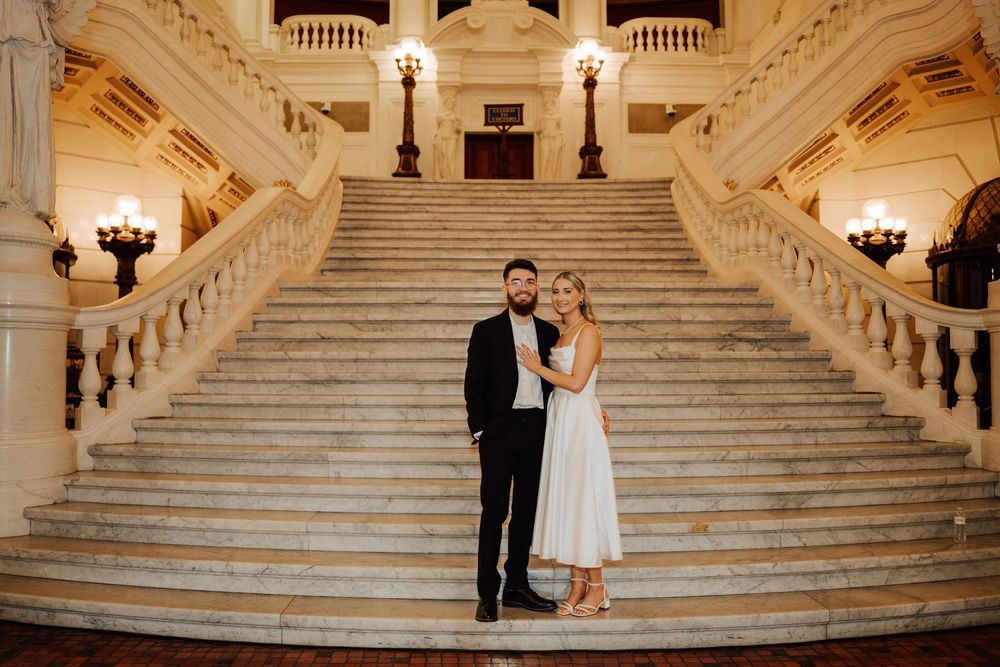 Couple poses on marble staircase in elegant grand hall, man in black suit, woman in white midi dress.