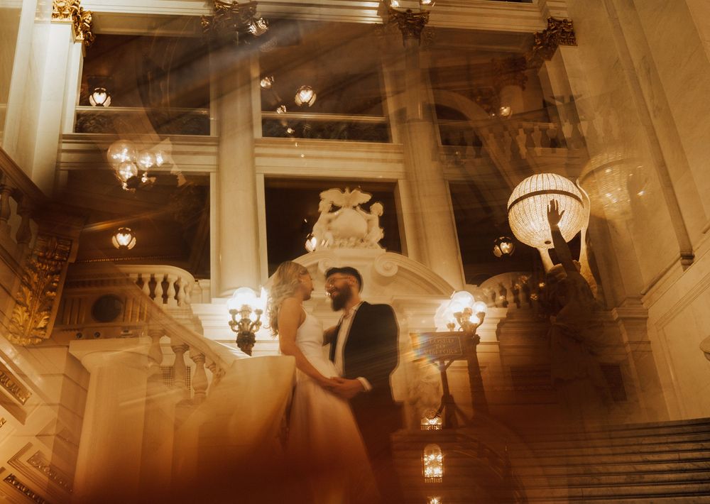 Elegant wedding couple embracing on grand staircase in ornate golden ballroom with chandeliers.