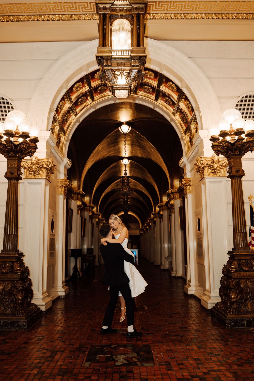 Couple embracing in an elegant ornate hallway with golden arches, columns, and warm lighting during a wedding celebration.