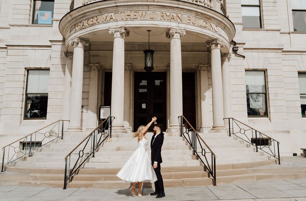 Bride and groom dancing on steps of elegant white stone building in wedding attire.