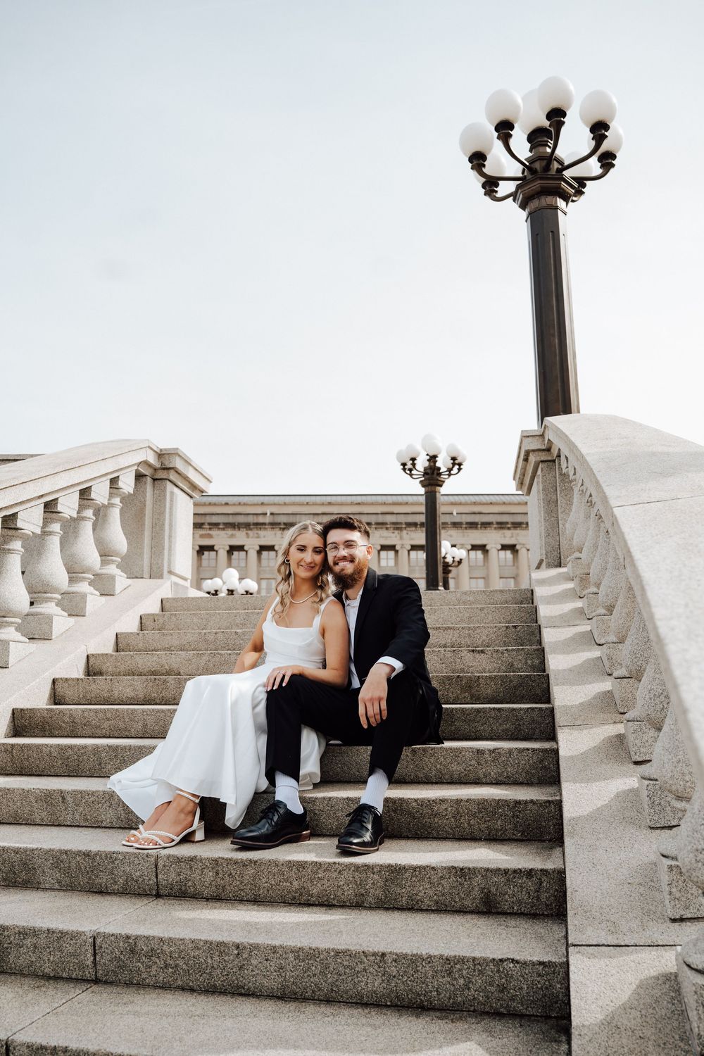 Bride and groom sitting together on grand stone steps with ornate lamp posts, smiling at camera on their wedding day.