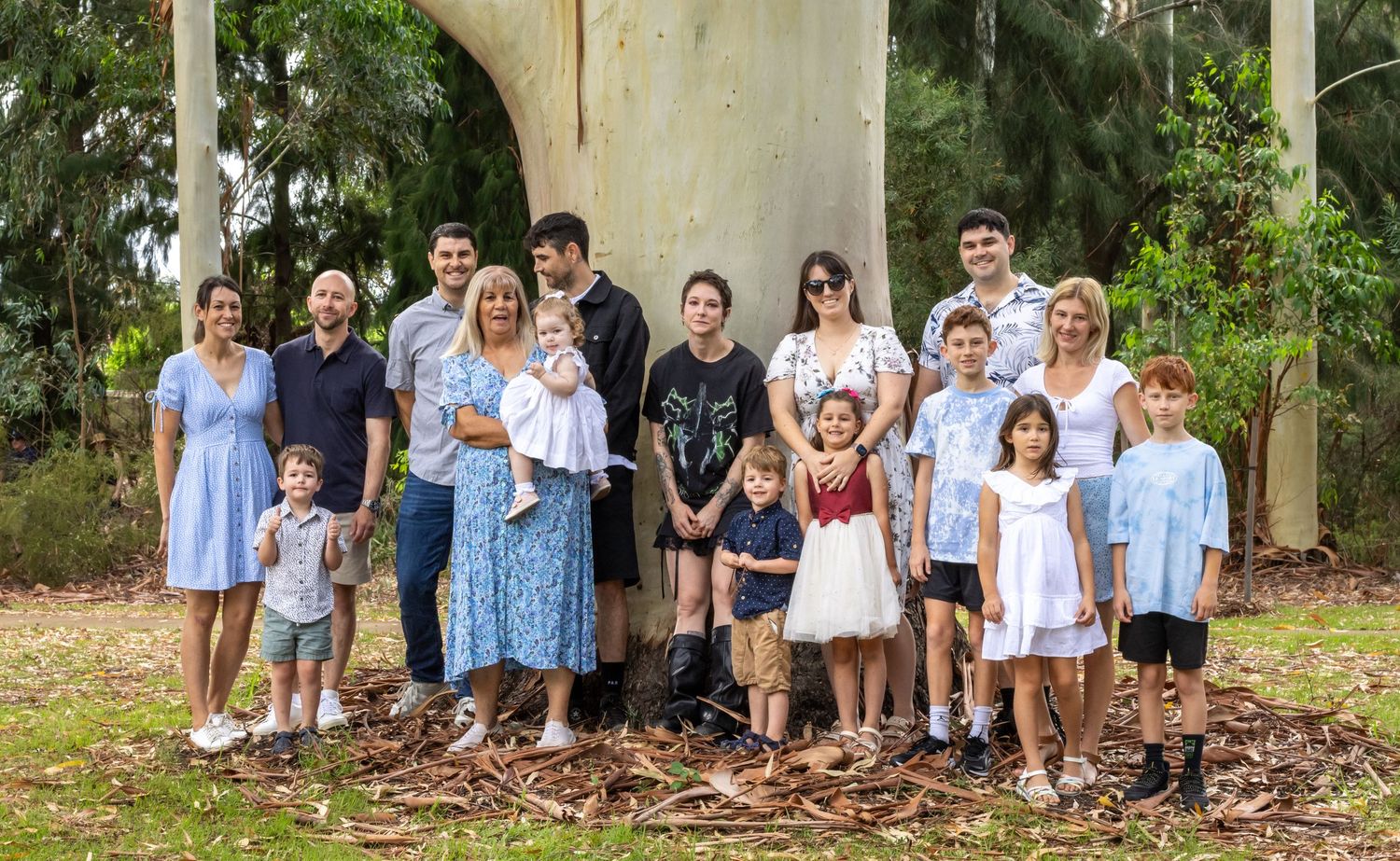 Large multigenerational family group portrait outdoors near a large tree in a lush green park setting.