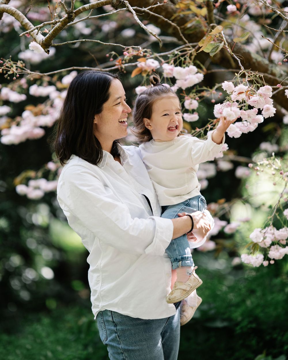 A woman in white shirt holds a toddler reaching for pink cherry blossoms in a lush garden setting.