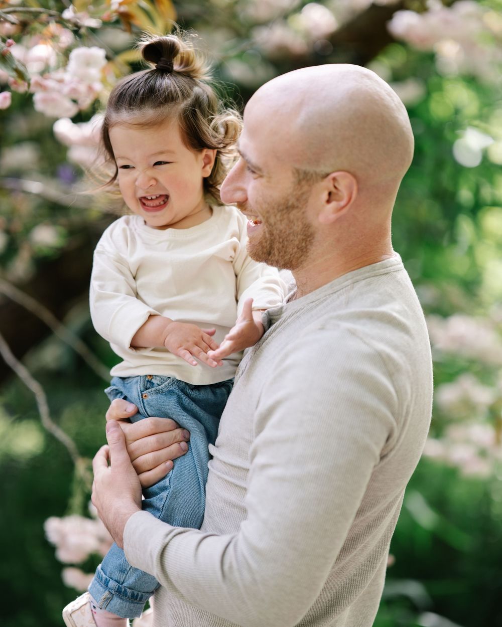 Bald bearded father holding laughing toddler girl among pink cherry blossoms in spring garden.