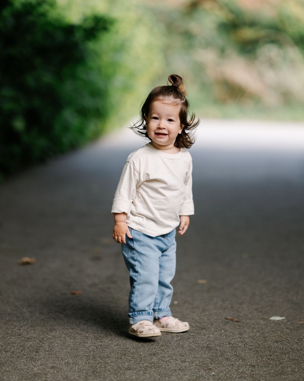 Toddler in white shirt and jeans smiling on a tree-lined path.