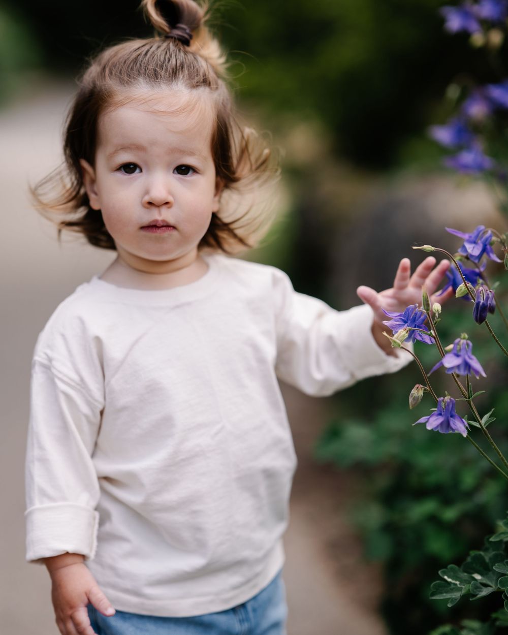 Curious toddler in cream sweater reaches toward purple wildflowers in a garden path.