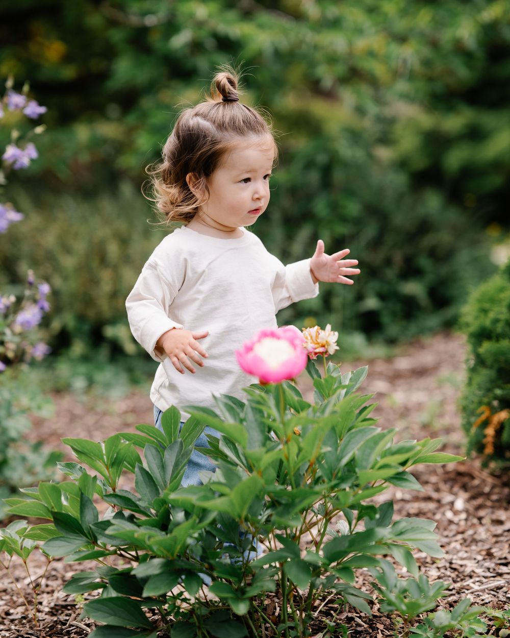 Toddler girl in white top reaching toward a pink peony flower in a lush garden setting.