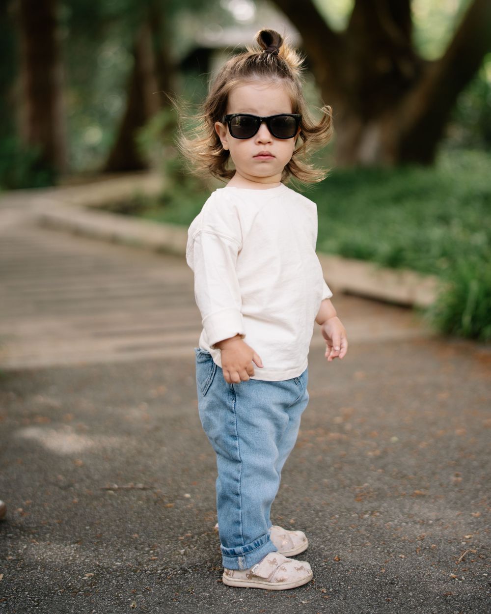Toddler in white shirt, jeans, and sunglasses standing on a path outdoors.