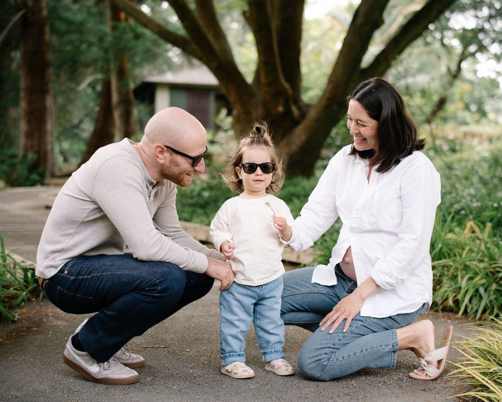Parents crouching beside toddler in sunglasses on garden path surrounded by lush greenery.