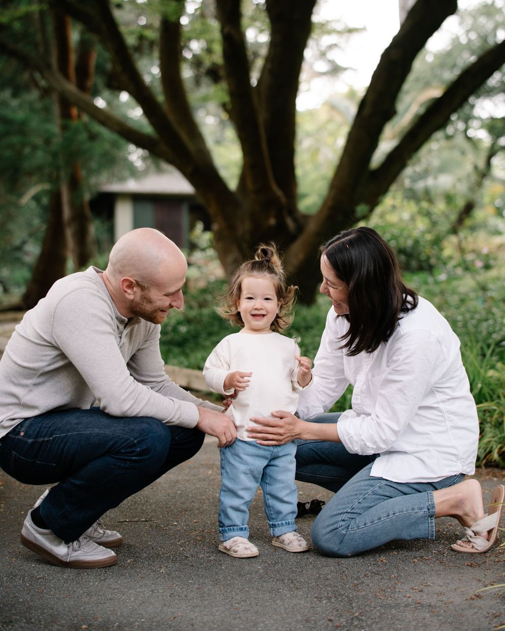 Happy family with toddler standing between smiling parents on outdoor path near large tree.