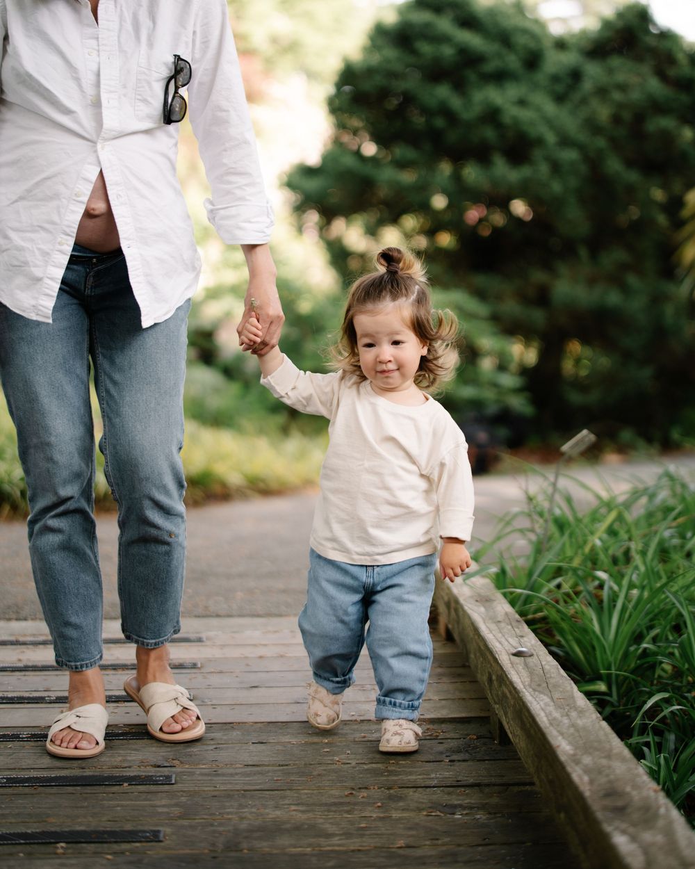 Toddler in cream shirt and jeans holds adult's hand while walking on wooden bridge path in lush garden.