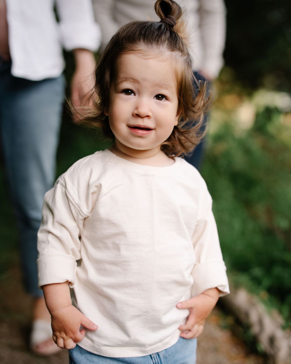 Adorable toddler girl in cream long-sleeve shirt and jeans smiles outdoors in natural setting.