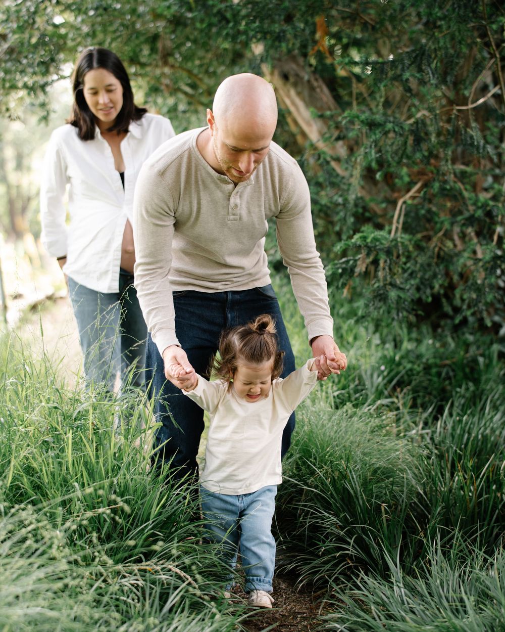 Parents guide their toddler daughter walking through lush green grass on a nature path.