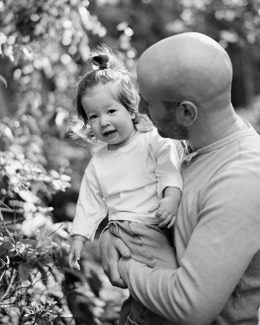 Black and white photo of a bald man holding a smiling toddler girl outdoors among lush green foliage.