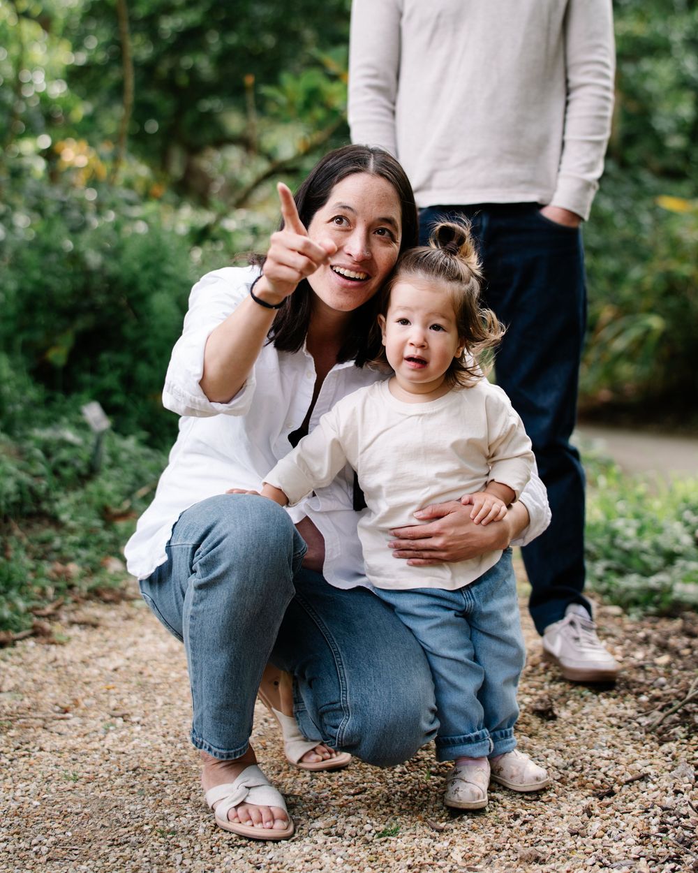 A smiling woman crouches with a toddler outdoors, pointing excitedly, both wearing casual white tops and jeans.