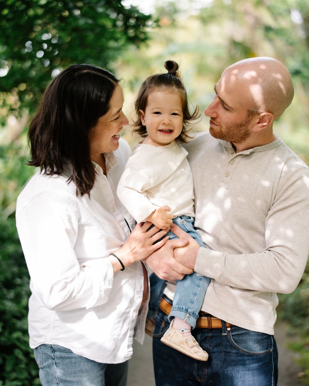 Smiling family with parents holding their toddler outdoors among green trees.
