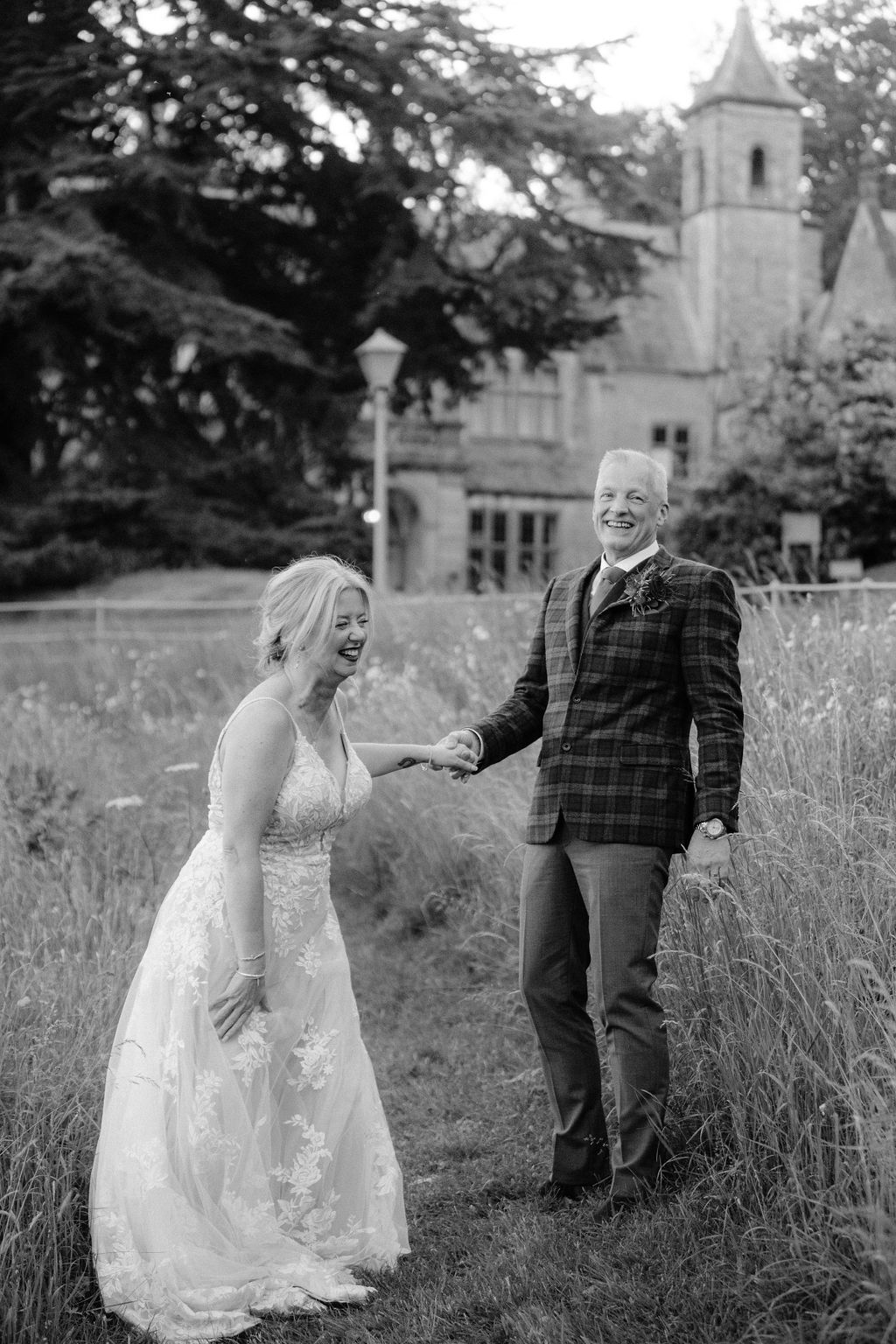 Black and white photo of a laughing bride in lace gown holding hands with groom in plaid suit outdoors.