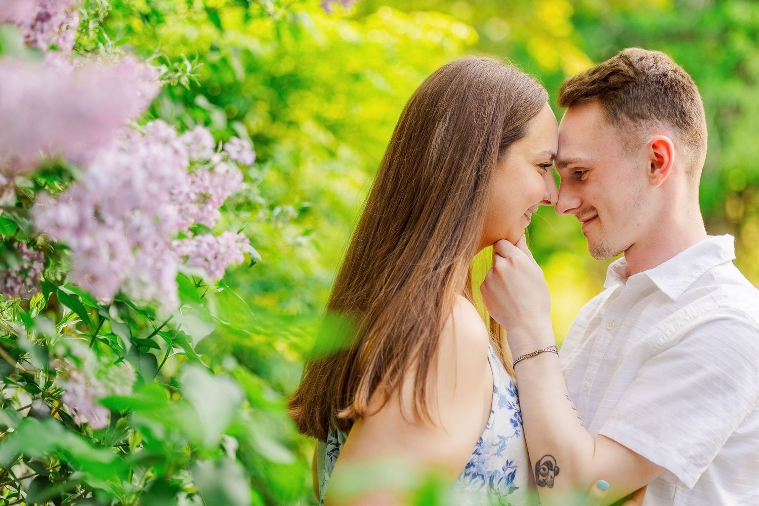 Couple touching foreheads surrounded by pink lilac blooms in a lush green garden.