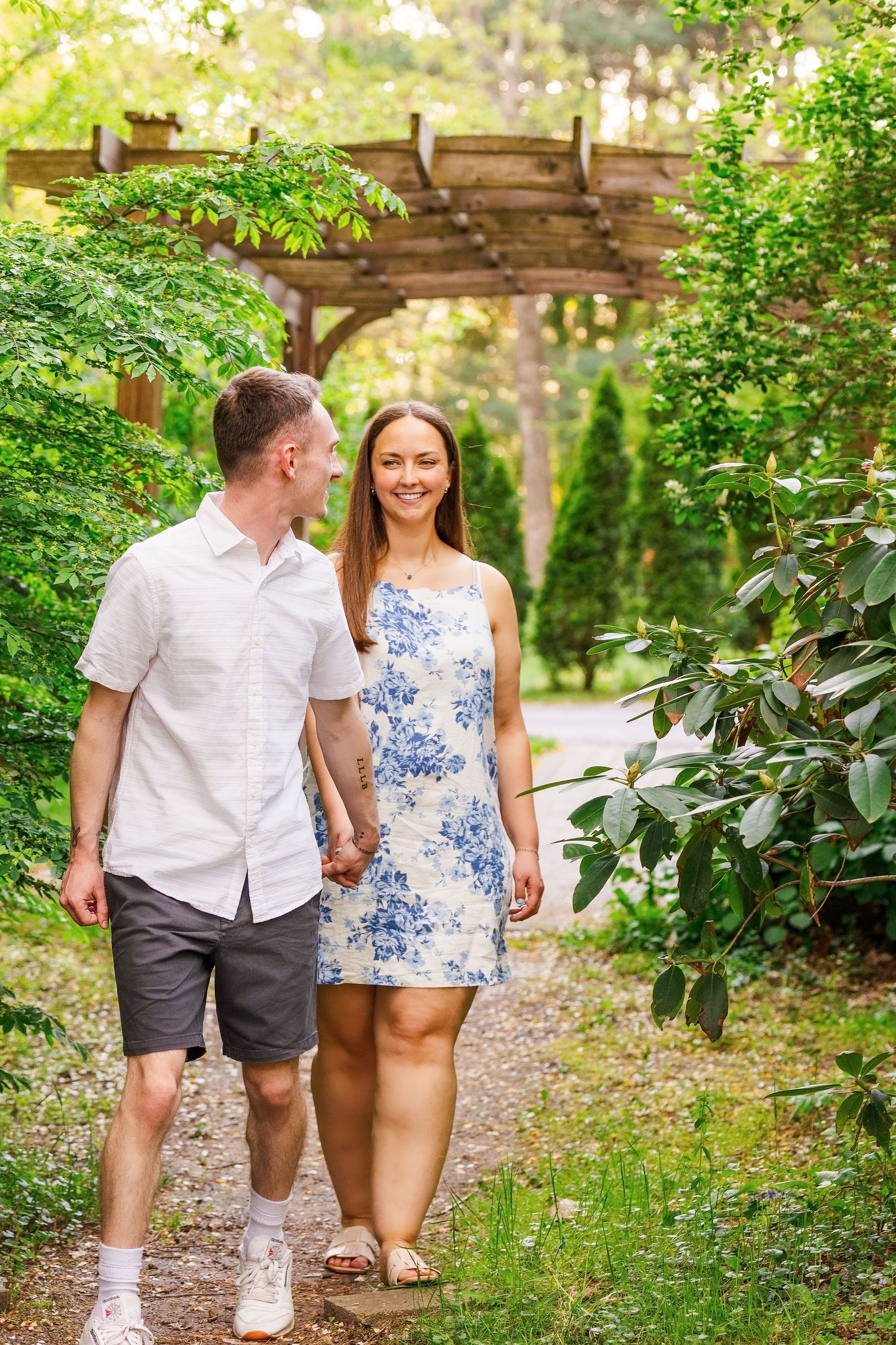 A smiling couple holds hands while walking through a lush green garden pathway with a wooden pergola.