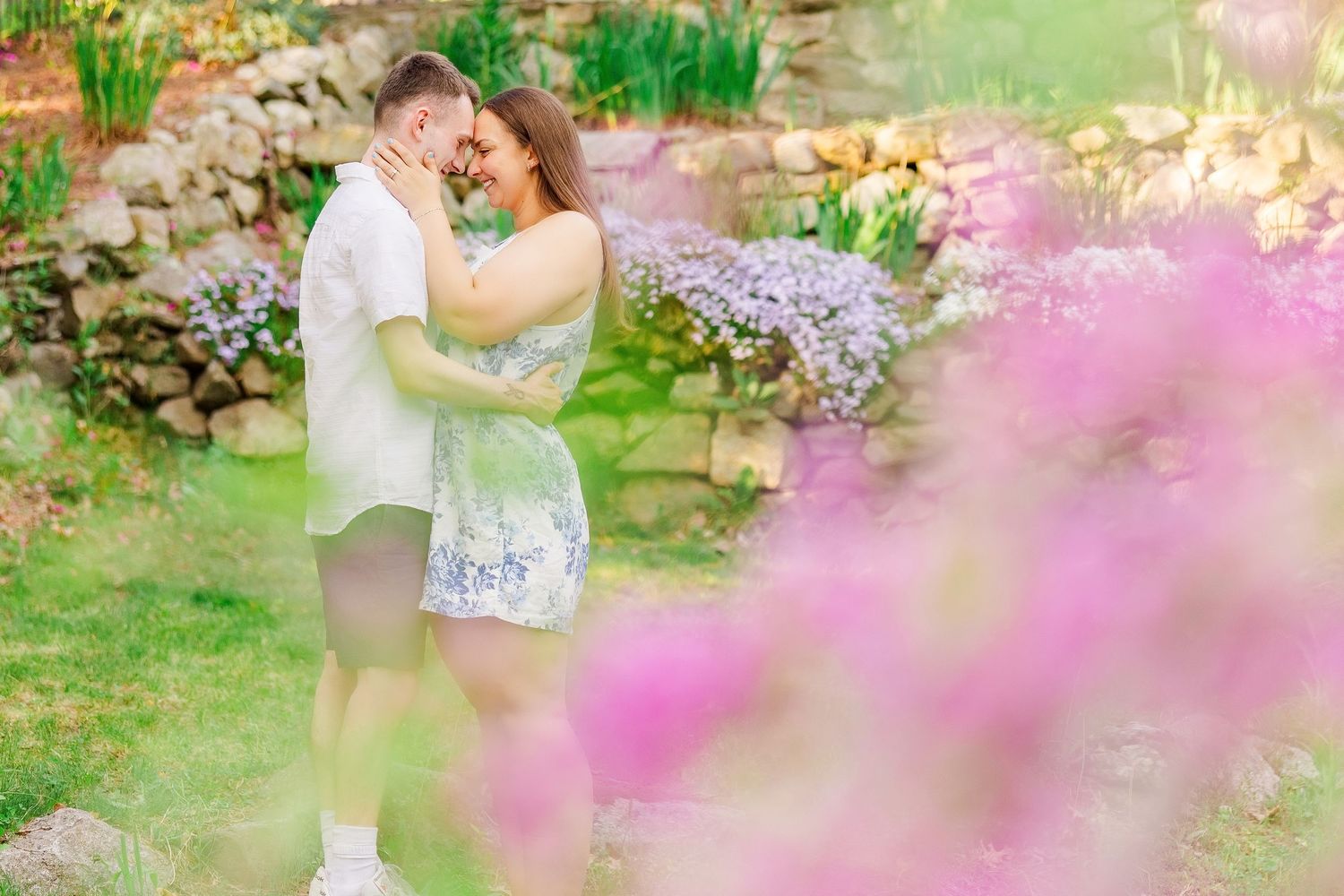 Couple embracing in a lush garden surrounded by vibrant pink and purple flowers during an outdoor engagement session.