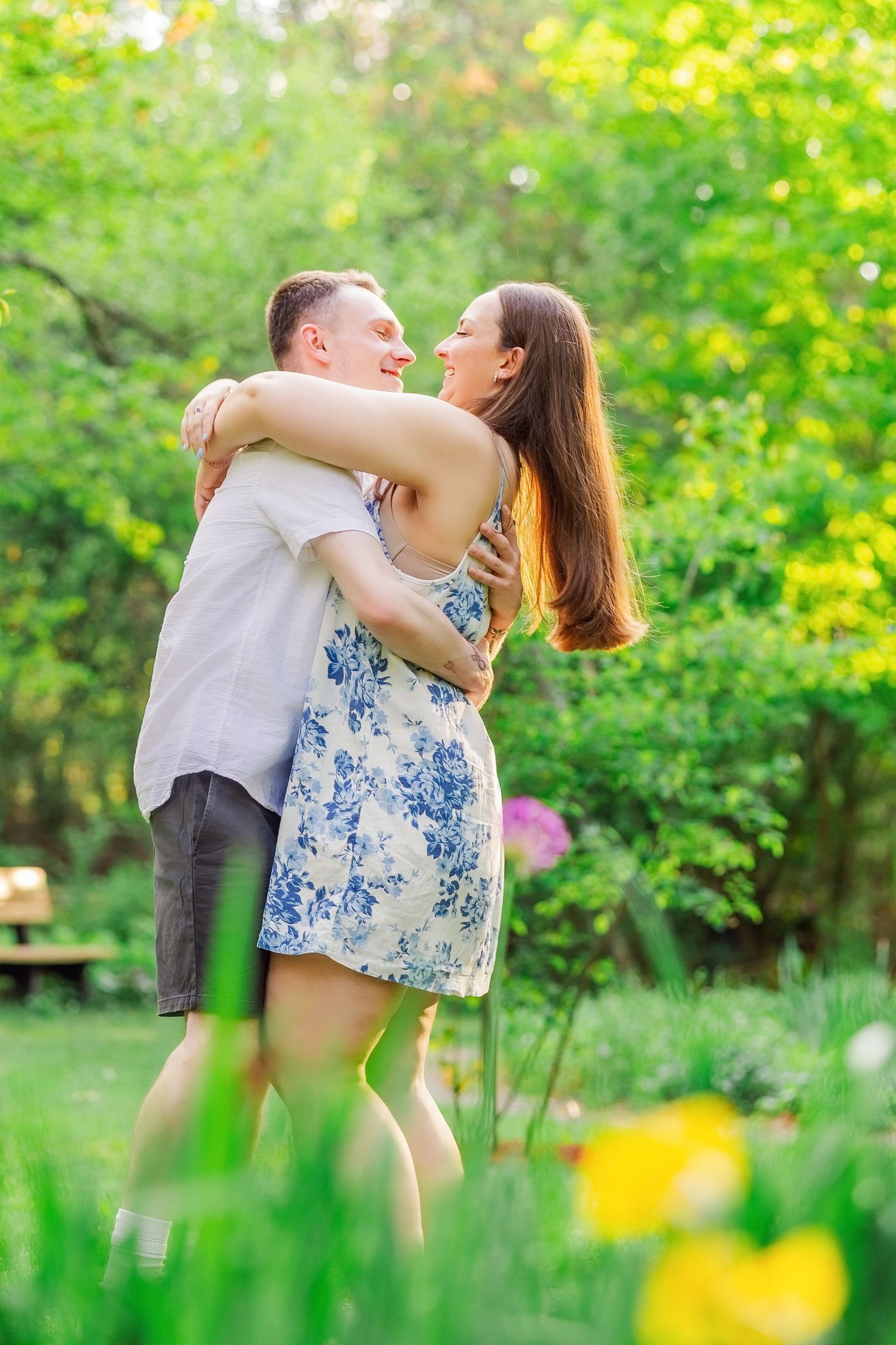 Happy couple embracing and laughing together in a sunny green garden, woman in floral dress lifted by man.