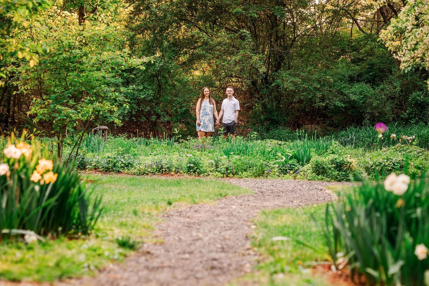 Couple holding hands walking through a lush green garden path surrounded by blooming spring flowers and tall trees.