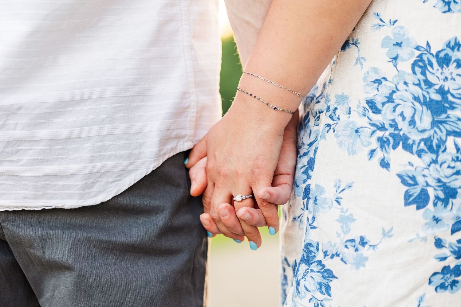 Couple holding hands outdoors, woman wearing floral dress and bracelet with engagement ring, man in white shirt and gray pants.