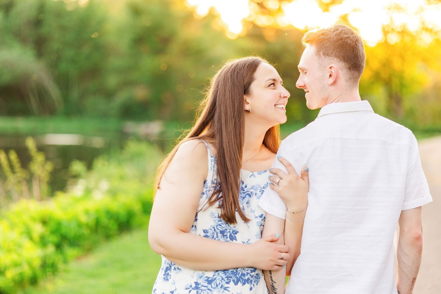 Smiling couple embracing at golden hour near a serene pond in a green park.