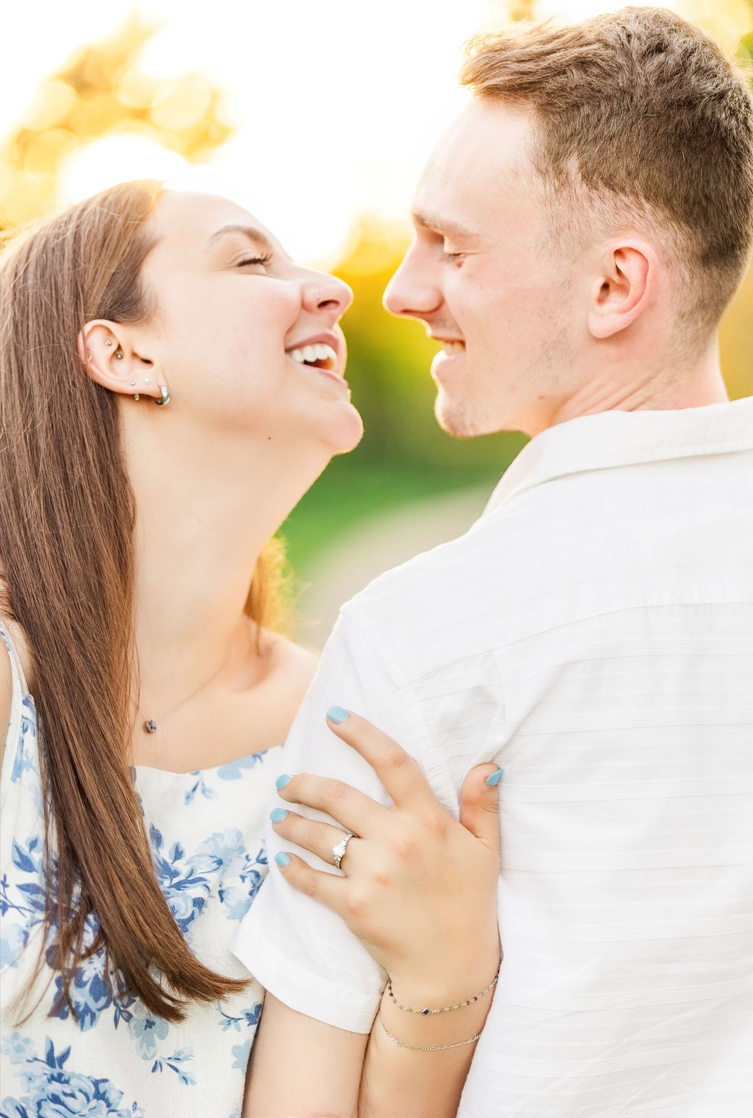 Happy couple sharing a joyful moment outdoors in golden sunlight, woman wearing floral dress and engagement ring.