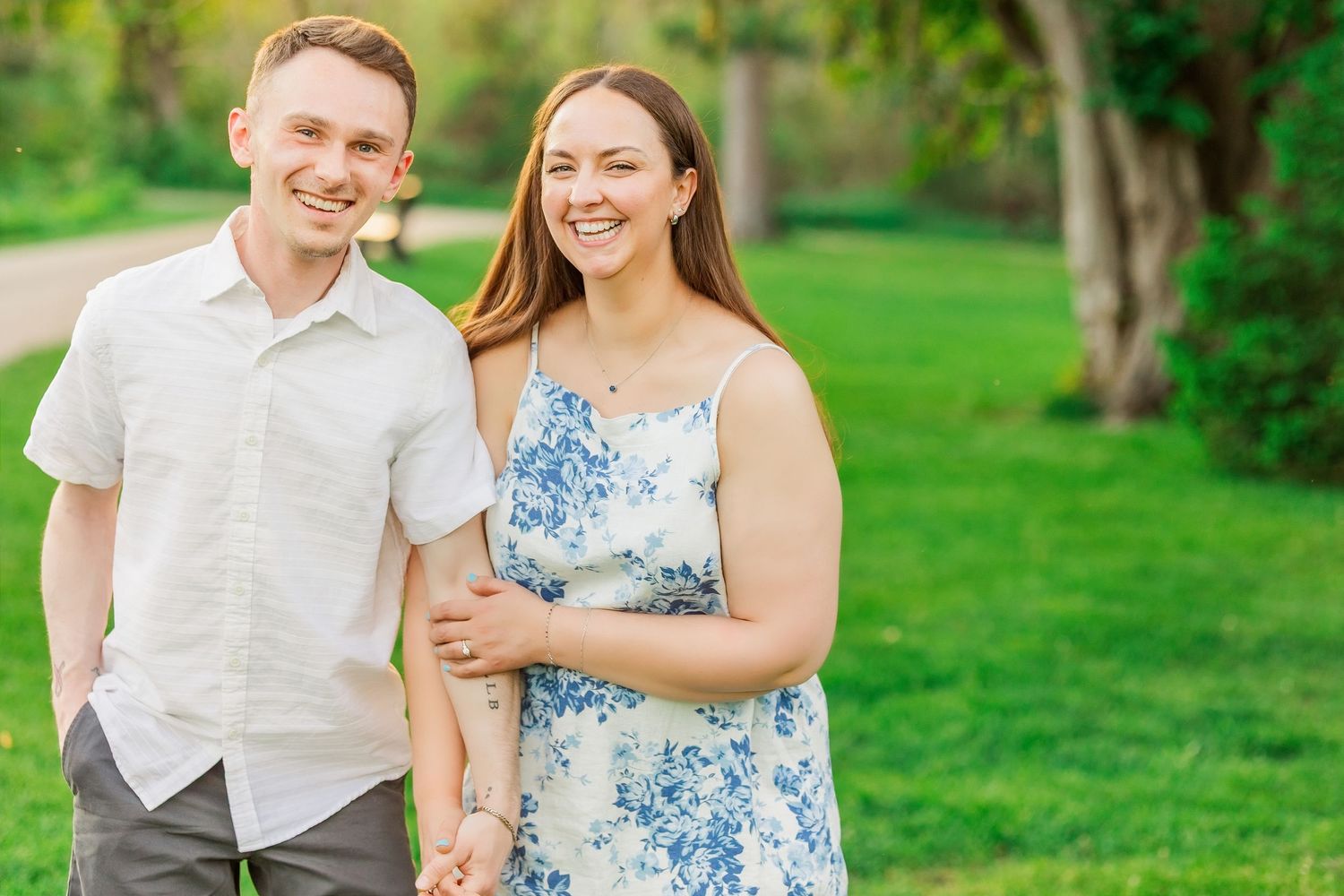 Smiling couple holding hands outdoors in a lush green park, man in white shirt and woman in floral dress.