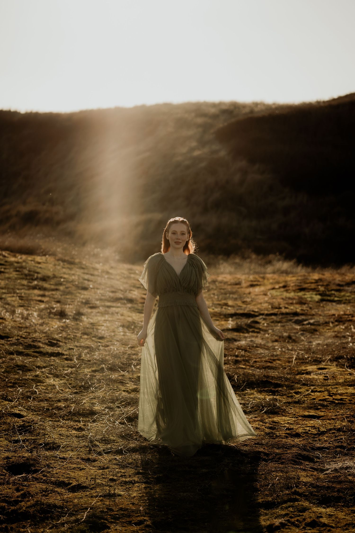 Black and white portrait of a woman in a flowing dress standing in tall grass, looking upward with hands clasped.
