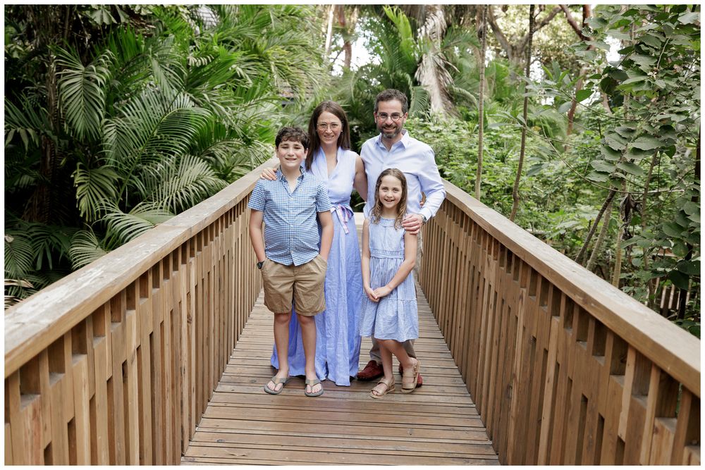 Family of four posing on a wooden bridge surrounded by lush tropical greenery, dressed in coordinating blue and white outfits.