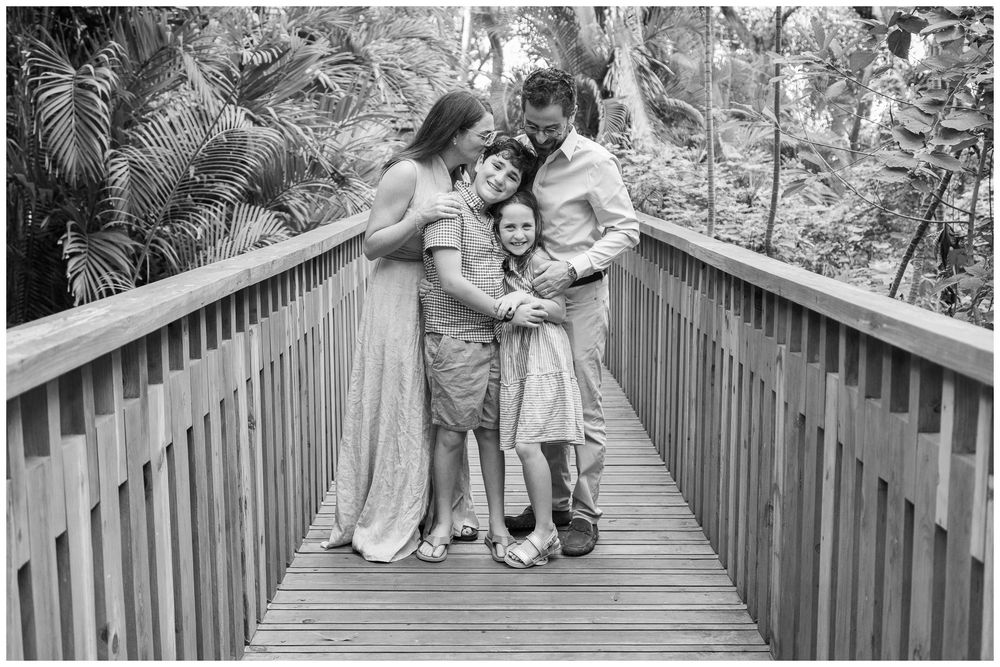 Black and white family portrait on wooden bridge surrounded by tropical foliage, parents embracing two children warmly.