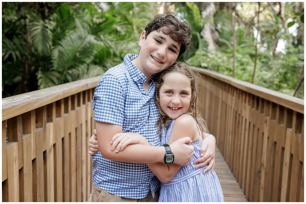 Two smiling children hugging on a wooden bridge surrounded by lush green trees in an outdoor portrait.